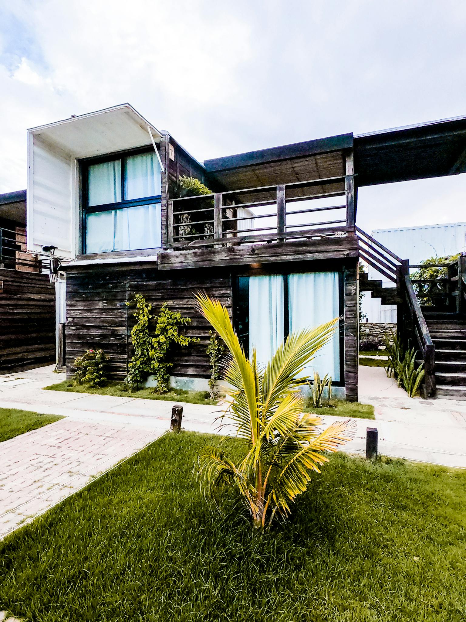 Contemporary two-story wooden house with lush greenery and palm plants.